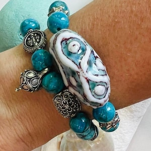 May include: A beaded bracelet featuring turquoise and silver-toned beads. The bracelet includes a large, white and blue patterned focal bead. Additional decorative silver beads add detail to the design. The bracelet is a piece of jewelry.
