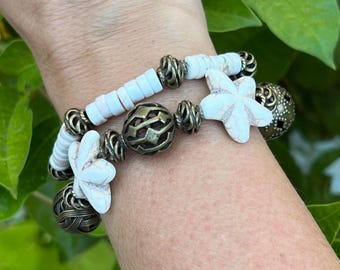 May include: A white and gold bracelet with white starfish charms and a large, ornate, gold bead. The bracelet is on a person's wrist.