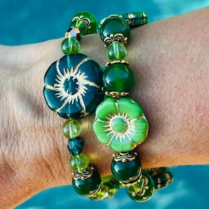 May include: Two beaded bracelets with green and gold accents. The bracelets feature large, dark green flower-shaped beads with gold detailing, interspersed with smaller, round green beads. The bracelets are on a wrist.