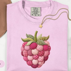 May include: A pale pink t-shirt featuring an appliqued raspberry design. The raspberry is crafted from diverse pink fabrics. A gold star pendant necklace is on the shirt. The tag reads "Comfort Colors".