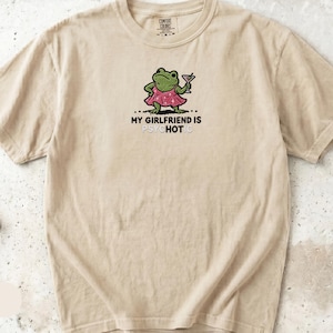 May include: An ivory short-sleeve t-shirt featuring a graphic of a green frog wearing a pink dress and holding a martini glass. Below the frog, the text reads MY GIRLFRIEND IS PSYCHOTIC, with the letters HOT highlighted in a different color.
