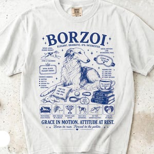 Puede incluir: Una camiseta blanca de manga corta con un estampado gráfico azul de un perro Borzoi rodeado de textos e ilustraciones que incluyen una taza de té, libros y varias etiquetas como "Elegant. Dramatic. 0% Interested." y "Grace in motion."