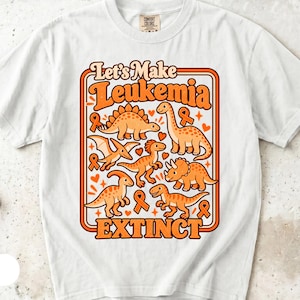 Könnte beinhalten: Ein weißes Kurzarm-T-Shirt mit einem Grafikdruck aus verschiedenen orangefarbenen Dinosauriern, kleinen Herzen und Awareness-Schleifen. Der Text Let's Make Leukemia EXTINCT steht in fetten orangefarbenen Buchstaben in einem rechteckigen Rahmen.