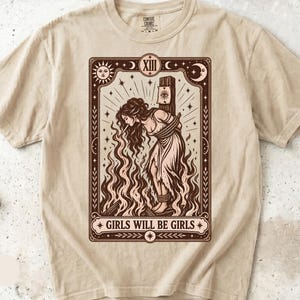 Puede incluir: Una camiseta de manga corta color marfil con un estampado gráfico marrón oscuro de una persona atada a un poste en medio de llamas. El diseño se asemeja a una carta del tarot con el número romano XIII y el texto GIRLS WILL BE GIRLS.