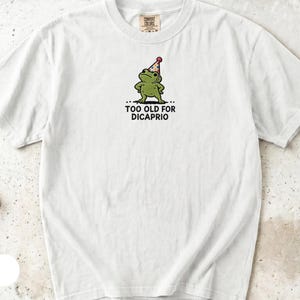 May include: A white short-sleeve t-shirt featuring a graphic of a green frog wearing a colorful polka-dot party hat. Below the frog, the text reads "TOO OLD FOR DICAPRIO" in black capital letters. The shirt has a crew neckline and a standard fit.