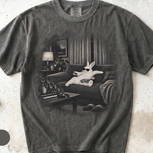 May include: A charcoal gray short-sleeve t-shirt featuring a graphic print of a white rabbit wearing sunglasses, lounging on a sofa while holding a remote control. The scene includes a lamp, a coffee table, and slippers, rendered in a monochromatic style.