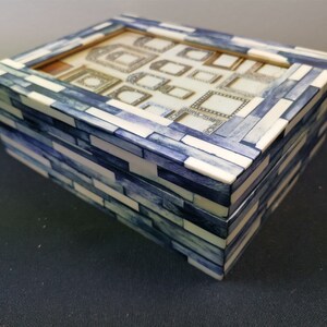 May include: A rectangular box featuring a mosaic pattern of alternating white and blue-dyed rectangular pieces. The lid has a glass window displaying a collection of small, varied decorative frames arranged in a grid on a light background.