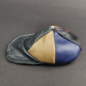 May include: A small, multi-colored leather cap featuring a patchwork design. The cap is constructed from individual panels of black, tan, and blue leather, with a curved brim and a rounded crown, showcasing a textured, stitched leather surface.