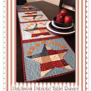 May include: Patchwork Patriotic Table Runner featuring a red, white, and blue star design with a blue background. The runner is made of fabric scraps and has a quilted texture. The runner is displayed on a wooden table with a bowl of red apples and a stack of plates.
