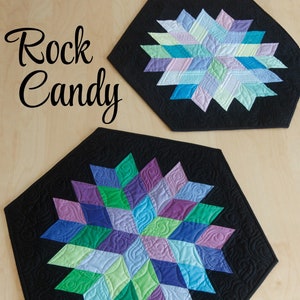 May include: Two quilted table runners with a geometric star design in shades of purple, blue, green, and white. The runners are black with a quilted pattern. The text "Rock Candy" is written in black cursive on a wooden surface. The text "little JAYBIRD QUILTS" is written in white on a black background.
