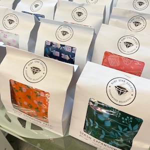 Peut inclure: Sacs en papier blancs avec une fenêtre transparente révélant des motifs de tissus colorés. Chaque sac porte un logo circulaire "RUBY STAR SOCIETY GUARANTEED DELIGHTFUL". Les sacs sont étiquetés "STASH EXPANSION PACK!"