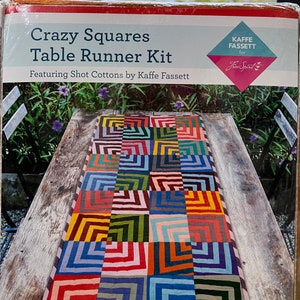 May include: A table runner kit featuring a colorful patchwork design with squares of various colors, including red, orange, yellow, green, blue, and purple. The kit is titled "Crazy Squares Table Runner Kit" and features "Shot Cottons by Kaffe Fassett" for Free Spirit.