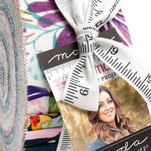 May include: A jelly roll of fabric strips featuring a variety of floral and botanical patterns in shades of purple, pink, orange, and dark blue. The roll is tied with a white ribbon printed with a black measuring tape design and a Moda Fabrics tag.
