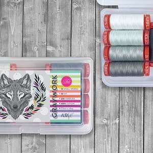 May include: Two clear plastic cases filled with spools of thread. One case features a wolf graphic and a label with text. The other case contains spools in shades of white, gray, and navy blue. The thread spools have red caps.