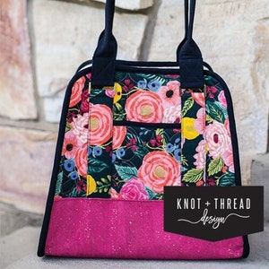 May include: A black and white floral print tote bag with a pink bottom and black handles. The bag has a large pocket on the front and the text "Knot + Thread design" is printed on the bottom right corner.