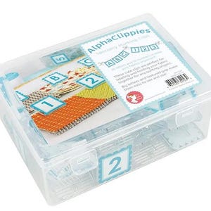 May include: A clear plastic storage box labeled "AlphaClippies" containing small, clear plastic clips. The box has a lid and a label with the letters "ABC" and numbers "123". The box also contains fabric samples with numbers and letters.