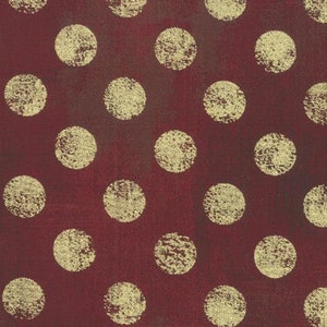 May include: Burgundy fabric featuring a repeating pattern of large, textured gold polka dots. The dots have a distressed, aged appearance. The fabric has a subtle, mottled texture. The bottom of the image shows a ruler with centimetre measurements.