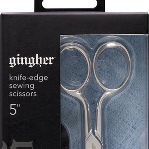 May include: A silver 5-inch Gingher knife-edge sewing scissors in a clear plastic package on a black background.