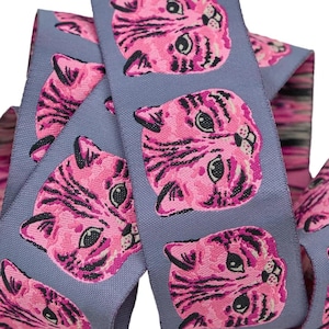 May include: A roll of gray ribbon featuring a repeating pattern of pink cat faces with black stripes. The ribbon is curled and layered, with the cat faces oriented in alternating directions. The ribbon's edges have a pink and black marbled design.