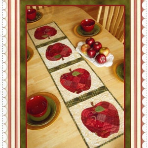 May include: A patchwork apple table runner with a red, green, and white color scheme. The runner is laid out on a wooden table with red bowls and apples on a white plate. The text "Patchwork Apple Table Runner" is at the bottom of the image.