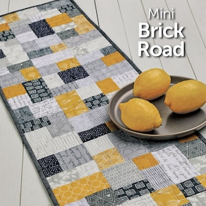 May include: A quilting pattern for a table runner called "Mini Brick Road" by Atkinson Designs. The pattern features a patchwork design with gray, white, and yellow squares. The pattern is shown on a table runner with lemons on a plate.