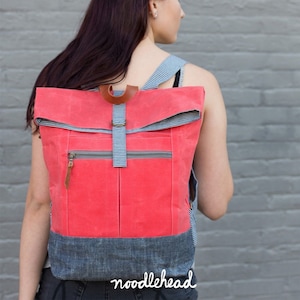 May include: A coral-colored foldover backpack with a zippered front pocket and a gingham patterned strap. The backpack is made of canvas and has a denim bottom. The text "noodlehead" is printed on the bottom of the backpack.