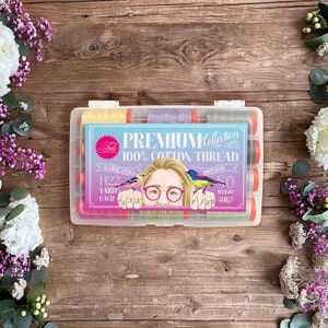 May include: A clear plastic storage box filled with 12 large spools of 100% cotton thread. The spools are variegated and come in a variety of colors. The box has a label that reads "PREMIUM Collection 100% COTTON THREAD" and includes details like "12 Large Spools", "1422 Yards Each", "Variegated", and "50 Weight".