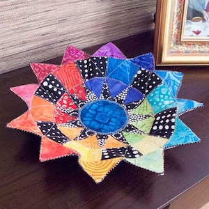 May include: A colorful patchwork fabric bowl with a starburst design. The bowl is made of various fabrics in shades of red, orange, yellow, green, blue, and purple. The center of the bowl is a blue fabric with a geometric pattern.
