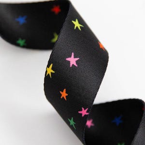 May include: Black ribbon featuring a repeating pattern of colourful stars in yellow, pink, orange, green, blue, and red. The ribbon is curled, showing the pattern on both sides. The background is white.