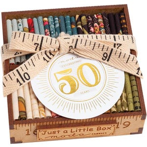 May include: A wooden box filled with fabric scraps, tied with a tan ribbon and a measuring tape. The box has a label that says "Just a Little Box moda FABRICS" and a circular label that says "moda CELEBRATING 50 YEARS".