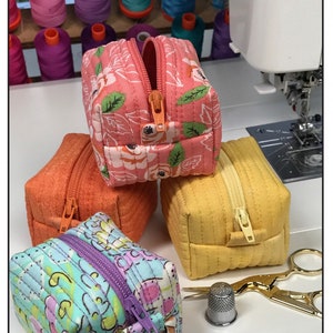 May include: Four small quilted zippered pouches in various colours and patterns, including pink with floral print, orange, yellow, and blue with a floral print. The pouches are stacked on top of each other. A thimble and gold scissors are in the foreground. The text "Bodobo Bags by Ticklegrass Designs" and "Bitsy Box" are visible in the image.