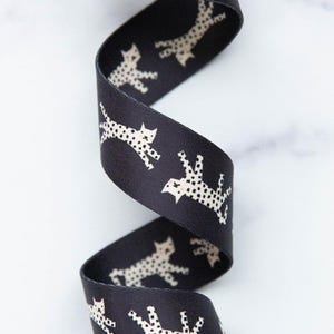 May include: A black grosgrain ribbon featuring a repeating pattern of stylized, off-white cats with black polka dots. The ribbon is coiled in a vertical spiral, showcasing the textured fabric surface and the consistent graphic cat print throughout.