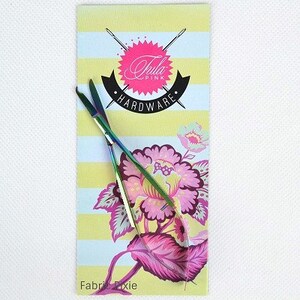 May include: A pair of rainbow-colored scissors rests on a decorative card. The card features a floral design in pink and purple, with the words "Tula Pink HARDWARE" and "Fabric Dixie". The background has yellow and blue stripes.