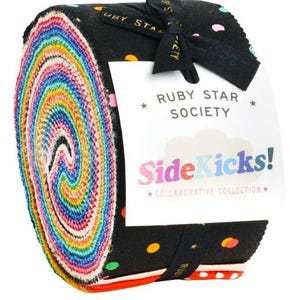 May include: A jelly roll of fabric strips featuring a vibrant rainbow gradient on the edges and a black fabric outer layer with colourful polka dots. A white paper label is tied around the roll with black ribbon, reading Ruby Star Society SideKicks!