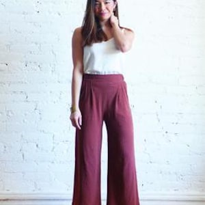 Peut inclure: Une femme porte un caraco blanc et un pantalon large bordeaux. Le pantalon a une taille haute et des plis. L'image comprend le texte "EMERSON PANT & SHORT" et "sewing pattern | sizes 0-18".
