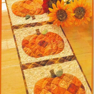May include: A rectangular quilted table runner featuring four distinct square panels, each displaying a patchwork pumpkin in shades of orange and brown. The background of each panel has a cream-colored swirling stitch pattern, and the runner is accented by three artificial sunflowers in red and yellow.