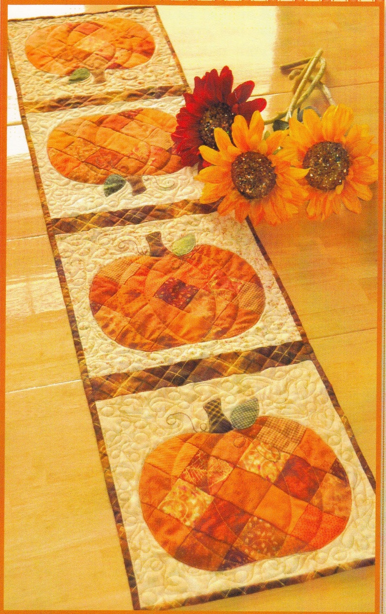 May include: A rectangular quilted table runner featuring four distinct square panels, each displaying a patchwork pumpkin in shades of orange and brown. The background of each panel has a cream-colored swirling stitch pattern, and the runner is accented by three artificial sunflowers in red and yellow.