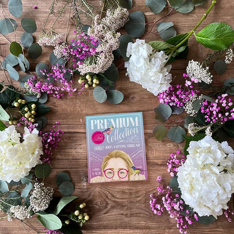 May include: A square package of Premium Collection 100% cotton thread featuring an illustration of a person with pink glasses and small birds. The label text includes 220 yards each and 50 weight. The package is surrounded by white hydrangeas.