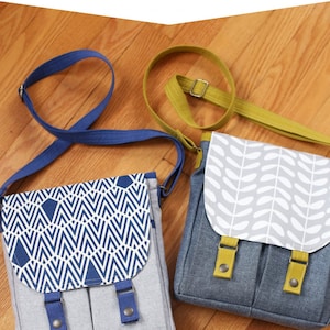May include: Two messenger bags with flap closures. One is gray with a blue and white geometric pattern, blue straps, and blue buckles. The other is gray with a white leaf pattern, yellow straps, and yellow buckles. The text "Campfire Messenger Bag" is at the top.