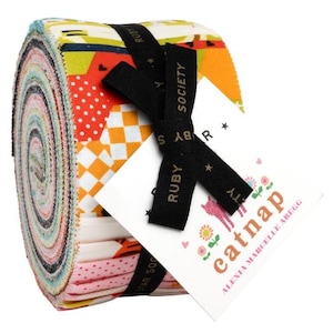 May include: A roll of colourful fabric with a black ribbon tied around it. The fabric is printed with various patterns, including polka dots, stripes, and geometric shapes. The ribbon has the text "Ruby Star Society" printed on it. A white card with the text "catnap" and "Alexia Marcelle Abegg" is attached to the ribbon.