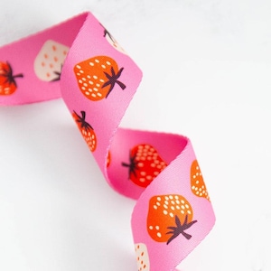 May include: Pink ribbon featuring a repeating pattern of strawberries in orange, red, and white. The strawberries have green stems and black seeds. The ribbon is curled and appears to be made of fabric.