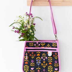May include: Black quilted crossbody purse with a floral pattern in yellow, orange, and purple. The purse has a pink trim and strap, and a tassel zipper pull. The text "Janet Mae Purse" is at the top.