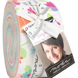 May include: Rolled fabric bundle with a floral pattern in pink, green, and orange on a white background. The fabric is tied with a white ribbon printed with measurements and the word "moda". A tag with a woman's photo and the "moda FABRICS + SUPPLIES" logo is attached.