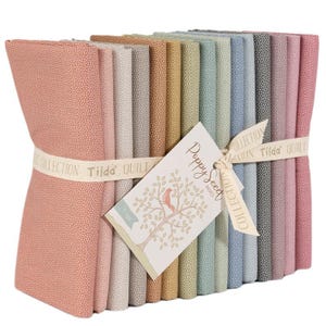 May include: A bundle of fabric squares in various pastel colors, including pink, gray, and blue, tied with a ribbon. The fabric has a small dot pattern. A tag with the words "Poppy Seed Basics" and a tree illustration is attached.