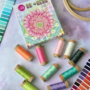 May include: A box of 80+ Neon embroidery floss spools in a variety of colors, including pink, orange, green, blue, and purple. The box features a floral design with a large pink flower. The spools are labeled "Aurifil" and "Made in Italy".