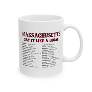 May include: White ceramic mug with the words "MASSACHUSETTS SAY IT LIKE A LOCAL" in red and black text. The mug lists phonetic pronunciations of Massachusetts town names. The mug has a curved handle.