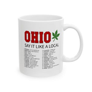 May include: White ceramic mug with the word "OHIO" in red block letters and a green leaf graphic. The mug features the phrase "SAY IT LIKE A LOCAL" and a list of Ohio place names with phonetic pronunciations.