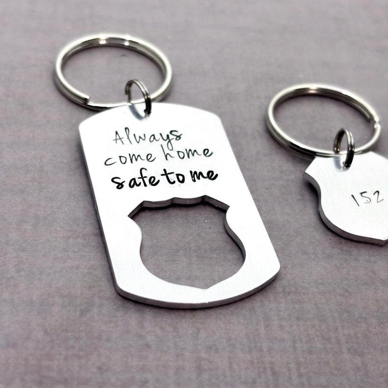 Two silver colored aluminum keychains with a police badge design. The larger keychain has the text 'Always come home safe to me' hand stamped on it. The smaller keychain is customizable.