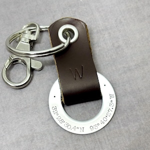 May include: A brown leather keychain with a silver ring engraved with the coordinates "32° 28' 30.4" N 93° 40' 27.3" W". The keychain has a silver split ring and a letter "W" embossed on the leather strap.