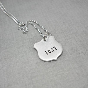 May include: A silver chain necklace with a silver shield pendant. The shield has the number "1027" engraved on it.